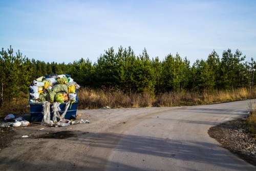 A large blue wheeled bin positioned at the side of a rural or semi-rural dirt road, filled with tightly packed yellow, black, and white plastic bags, along with loose rubbish spilling onto the ground nearby. The bin's lid is open, revealing its contents, and debris such as paper and plastic waste can be seen around its base. The background features a dense line of green foliage and trees under a clear, pale blue sky, indicating an outdoor environment likely in the vicinity of Leyton or nearby Essex. The scene suggests waste collection or disposal, relevant to rubbish removal services offered by Garden Clearance Leyton, situated in the London borough of Waltham Forest postcode area, with the setting emphasizing the importance of proper rubbish handling in residential or semi-rural areas.