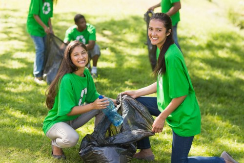 A group of young volunteers wearing bright green t-shirts with recycling symbols are participating in a community rubbish cleanup in a grassy park area. The focal point shows two smiling women, one kneeling and the other standing, as they hold a large black garbage bag filled with collected waste. The woman kneeling has long dark hair, beige trousers, and is holding a plastic bottle, while the standing woman has long dark hair and blue jeans, actively engaged in the collection process. In the background, other volunteers are visible, some holding additional black rubbish bags and wearing similar green shirts, all working together to help clear litter from the outdoor environment. The scene is well-lit with natural daylight, conveying a positive and proactive approach to rubbish removal and waste management in a community setting, consistent with services like those offered by Garden Clearance Leyton related to rubbish collection and environmental cleanup within the Leyton area.