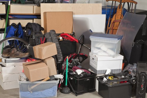 A large collection of assorted waste and household rubbish stacked together indoors, including cardboard boxes, plastic storage containers, black and grey rubbish bins, a vacuum cleaner, and various discarded items such as tools, fabric, and packaging materials. The background features a metallic chair, stacked shelves with books and storage boxes, and a cluttered environment typical of a storage or clearance setting. The items are placed on a concrete floor, with some leaning against each other and the surrounding environment. The scene illustrates typical household or garden clearance waste that Garden Clearance Leyton might collect, emphasizing the presence of multiple materials like cardboard, plastic, and metal objects associated with domestic clutter or debris.