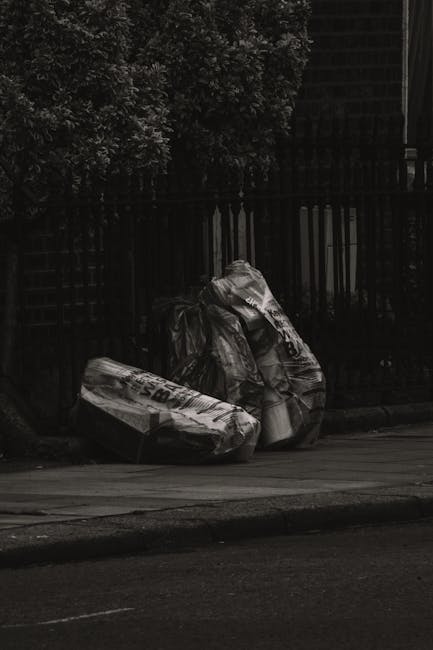 Three large black plastic rubbish bags filled with waste are placed on a paved sidewalk next to a black metal fence, which separates the sidewalk from a garden or yard area with dense trees and bushes
