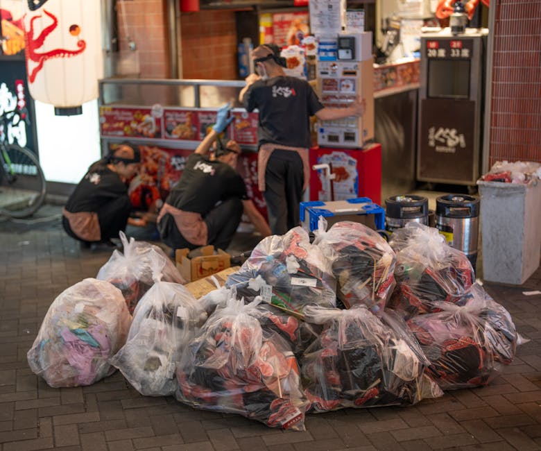 A person in a camouflage-patterned jacket is seen lifting a large, irregularly shaped bag of waste on a city street at night. The bag appears to be made of thick, durable plastic with a slightly crink
