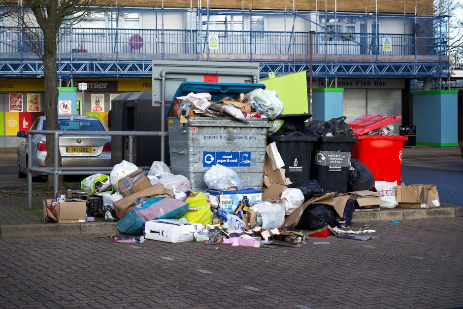 Two large black plastic bags filled with rubbish, positioned on the pavement next to a black metal fence and a brick wall, with some crumpled paper and cardboard visible on the bags' surfaces. The sce