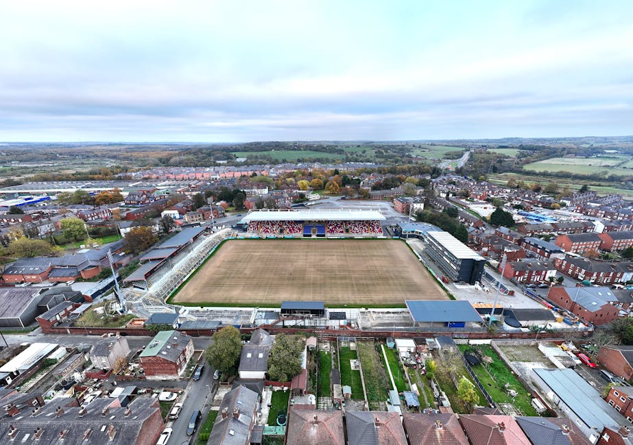 An aerial view of a football stadium situated within a residential and commercial area, with surrounding houses, gardens, and streets. The stadium features a central grass pitch that appears to be und